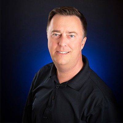 The image features a man smiling at the camera, standing against a dark background with a blue tint.