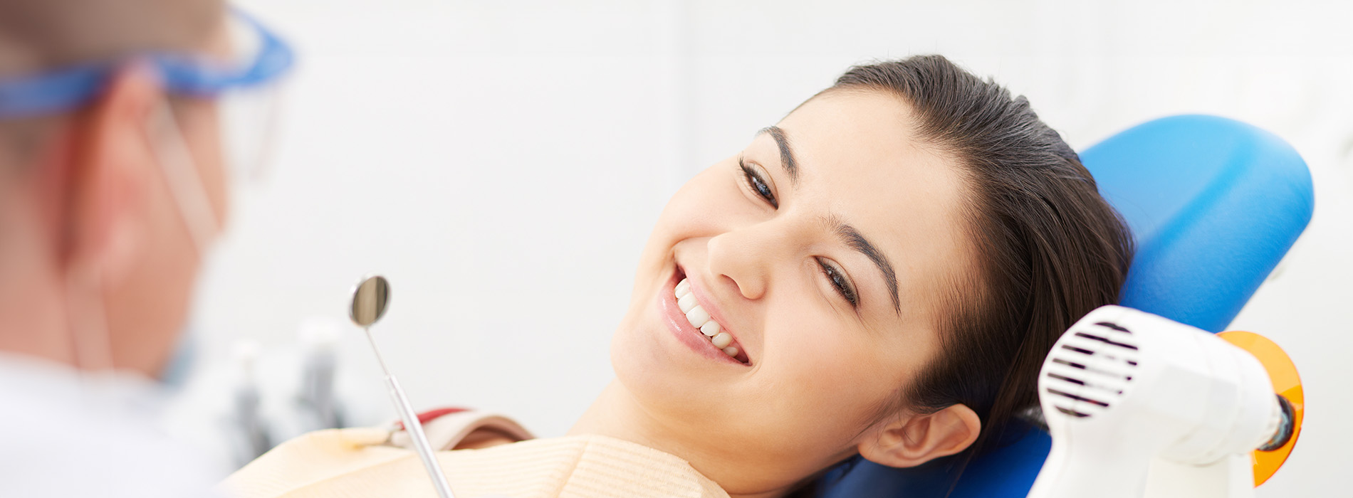 The image shows a woman sitting in a dental chair with her head turned towards the camera, smiling at the viewer, while receiving dental care, with a dentist visible behind her.