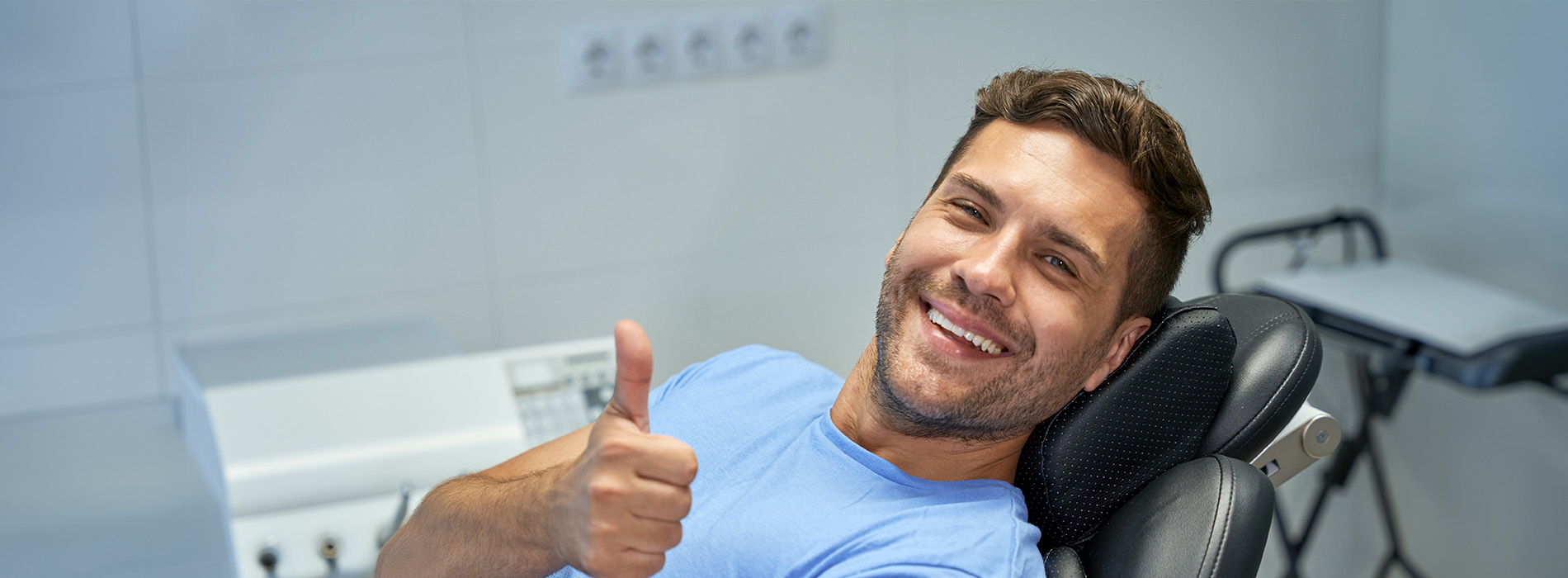 The image shows a man sitting in a dental chair with his thumbs up, smiling towards the camera.