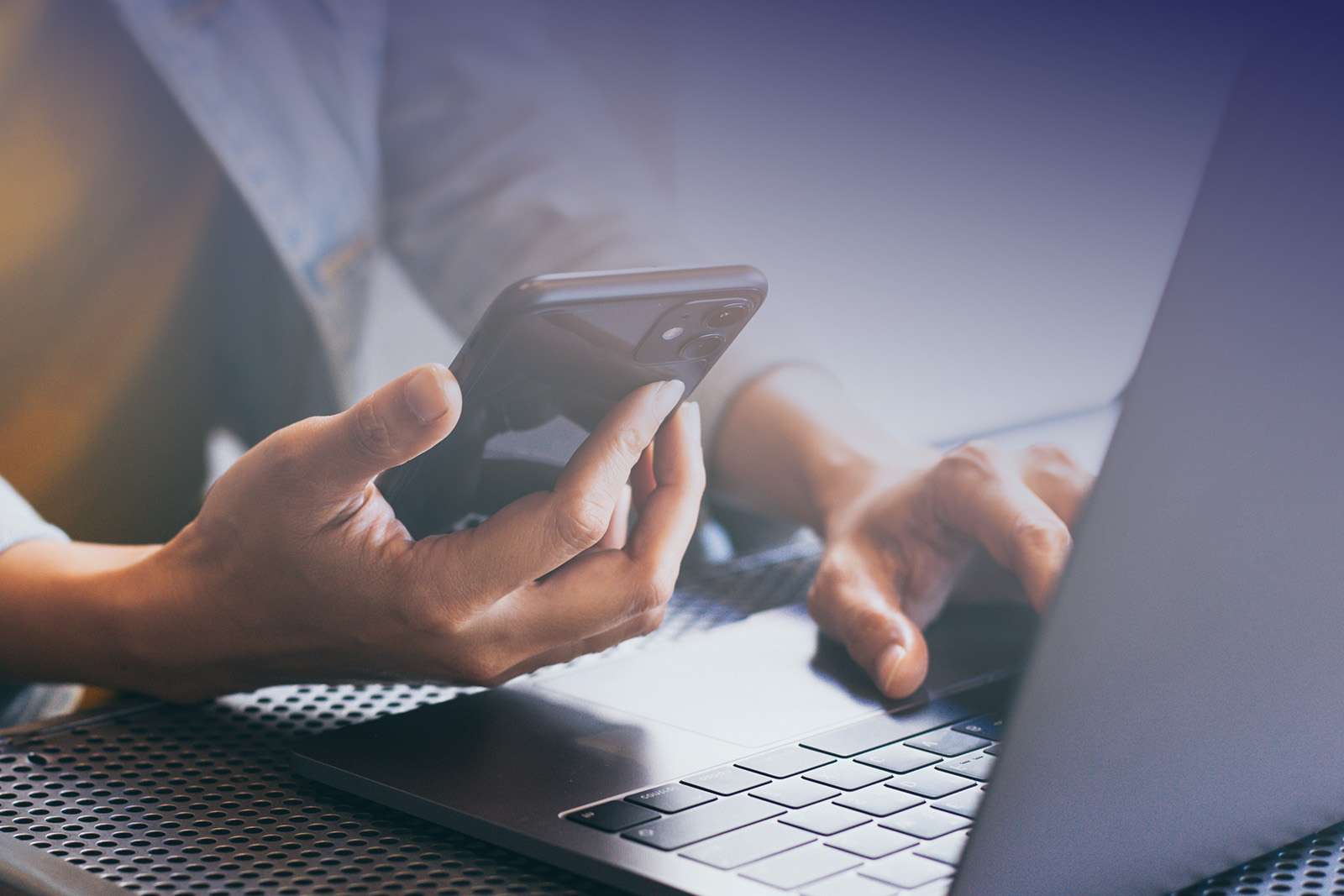 The image shows a person s hands holding a smartphone while sitting at a table with a laptop open in front of them.
