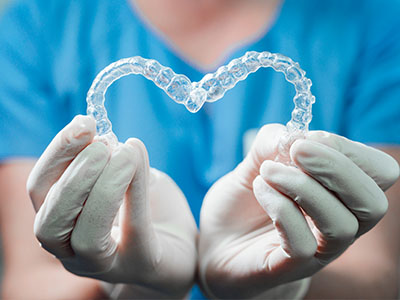 The image shows a person s hands holding a transparent heart-shaped object with a clear plastic texture, possibly a dental appliance, against a blue background.