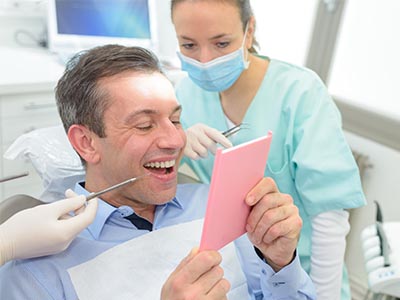The image shows a man sitting in a dental chair with his mouth open, holding up a pink card with a smile on his face, while a woman wearing a surgical mask stands behind him with a tablet displaying a digital image of the same card.