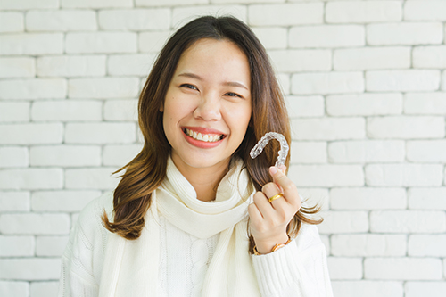 A woman with a radiant smile holds a white sign with a smiley face symbol, set against a brick wall background.