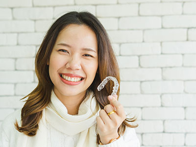 The image shows a smiling woman holding a toothbrush with her right hand, standing against a brick wall background.
